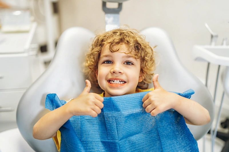 A cheerful young boy with curly hair giving two thumbs up while wearing a blue bib in a dental chair.