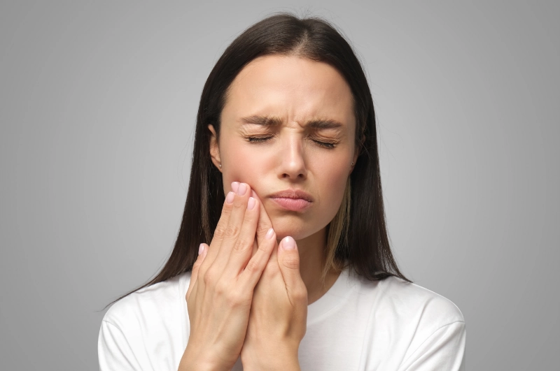 A young woman holding her cheek with both hands and wincing in pain from a toothache.
