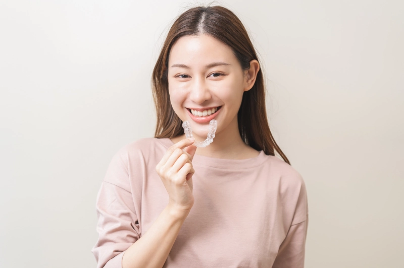 A smiling young woman holding up a clear plastic aligner tray in front of her smile.